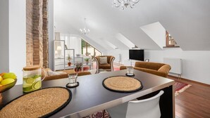 A modern loft-style living room with a dining area, featuring exposed brick walls, a chandelier, and a large window providing natural light. - Garni Hotel VIRGO (Bratislava)