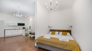 A modern hotel room with a wooden floor, a bed with a mustard-colored blanket, a wooden headboard, a white bedside table, a white chair, a small table with a vase of flowers, a white cabinet, a microwave, a flat-screen TV, and a chandelier. - Garni Hotel VIRGO (Bratislava)