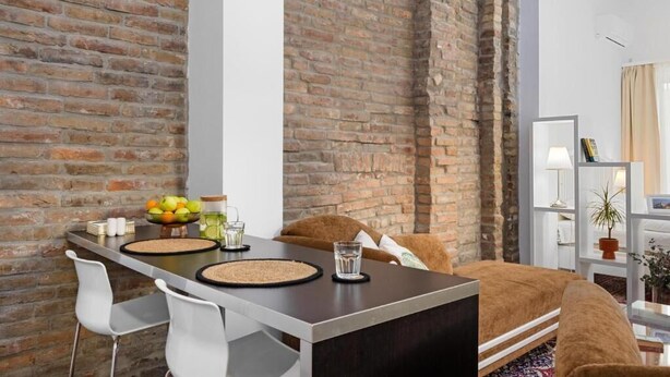 A modern room with exposed brick walls, a dark wooden desk, white chairs, a brown sofa, and a white shelving unit with a lamp and a plant. - Garni Hotel VIRGO (Bratislava)