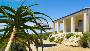 Una casa con fachada blanca, un patio con techo de paja y vista al mar.
