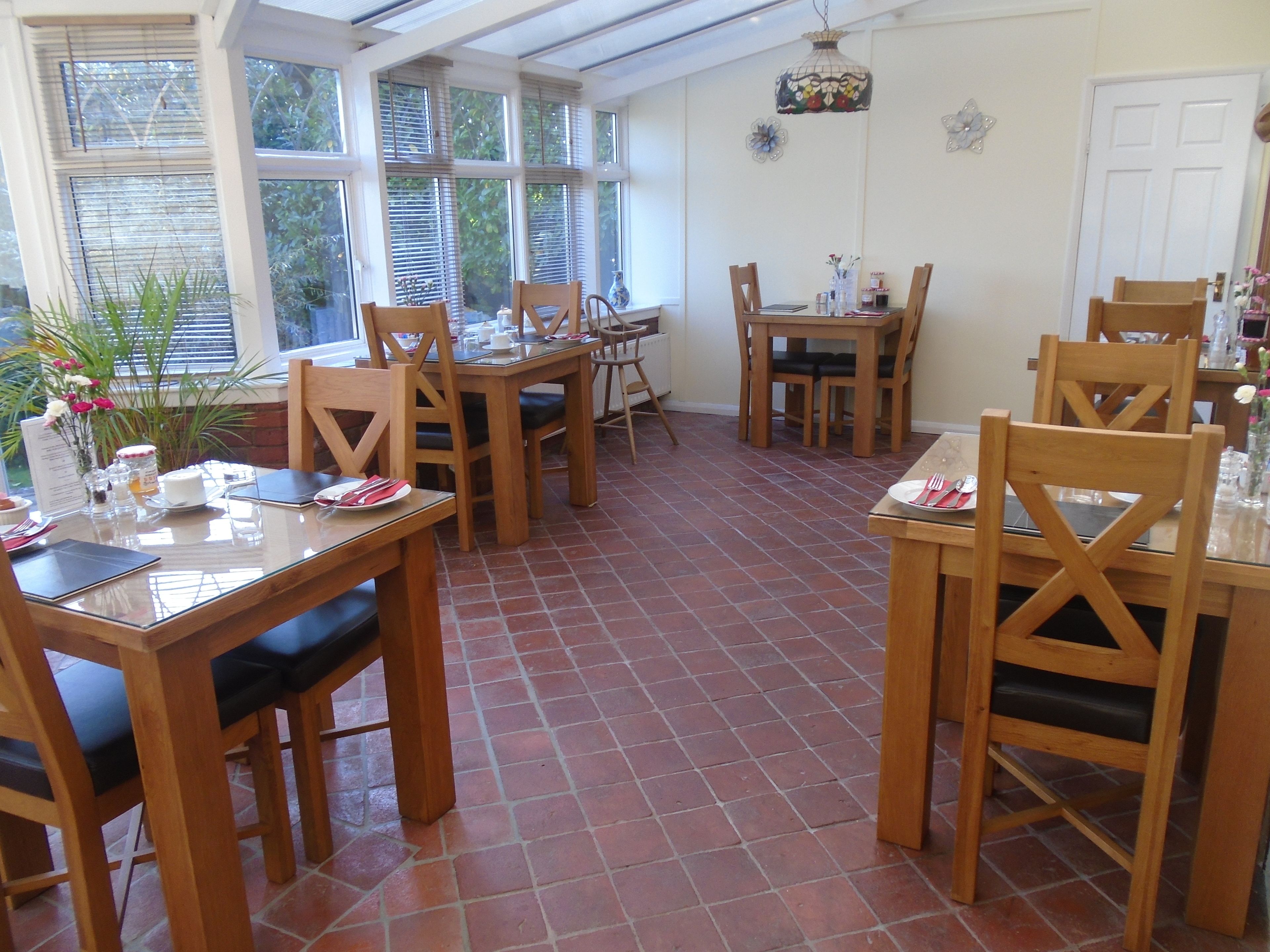 A dining area with wooden tables and chairs, a tiled floor, and large windows with blinds.