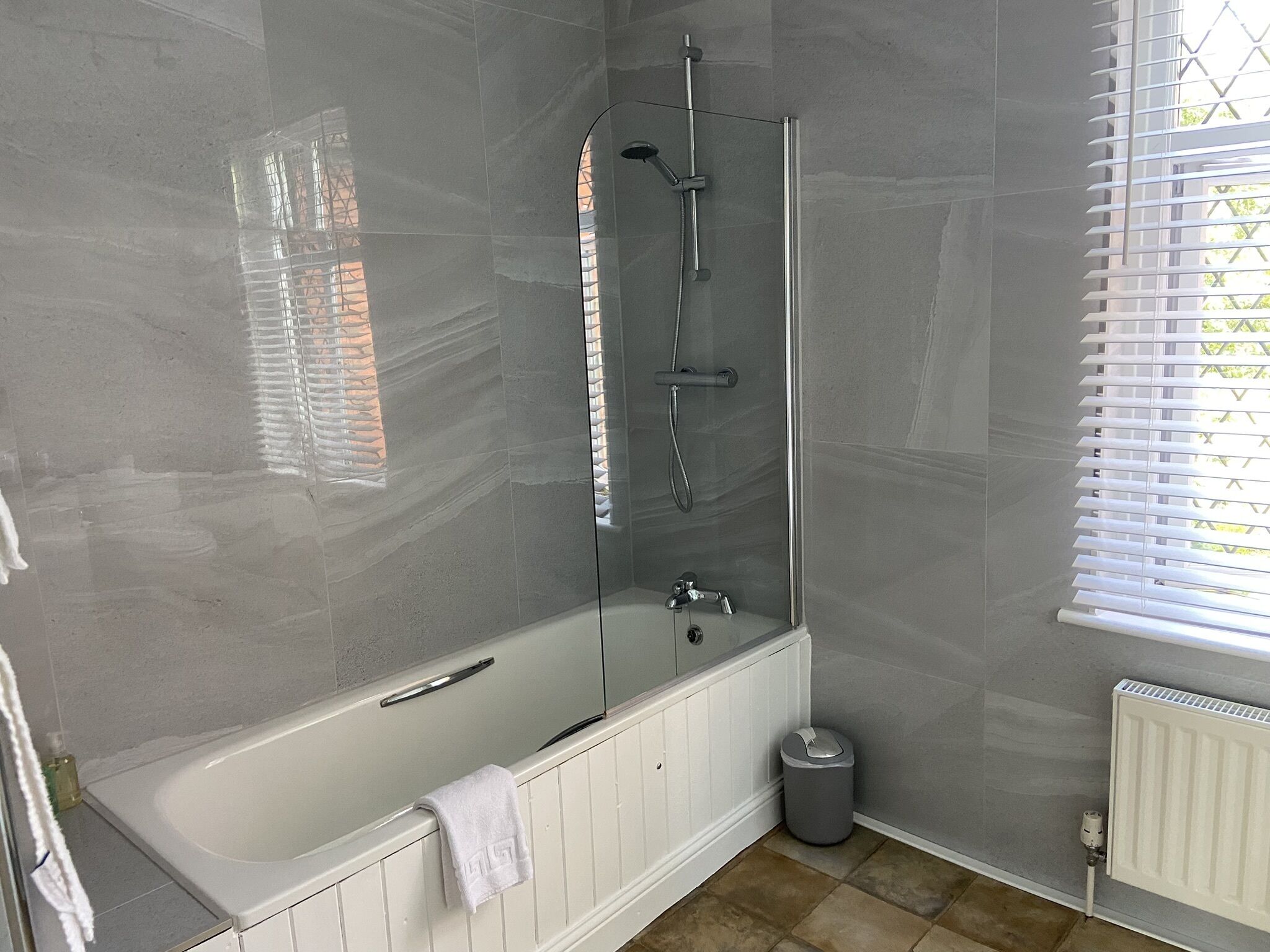 A modern bathroom with a glass-enclosed shower, a white bathtub, and a window with blinds.