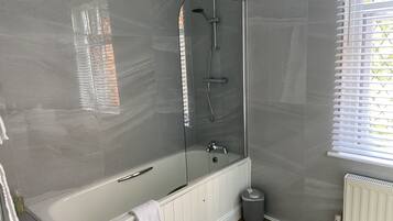 A modern bathroom with a glass-enclosed shower, a white bathtub, and a window with blinds.