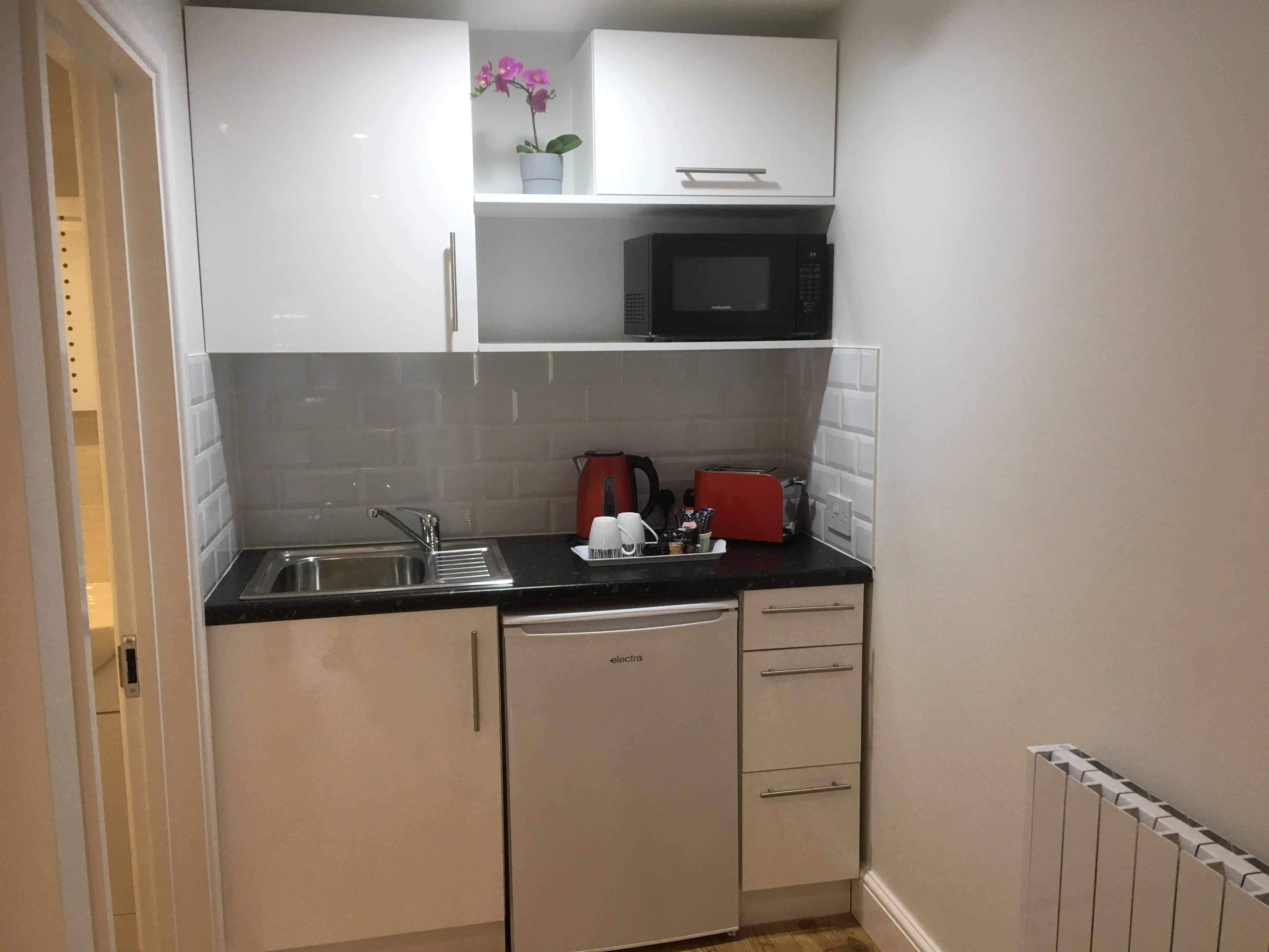 A compact kitchen with white cabinets, a black countertop, a microwave, a red kettle, and a dishwasher.