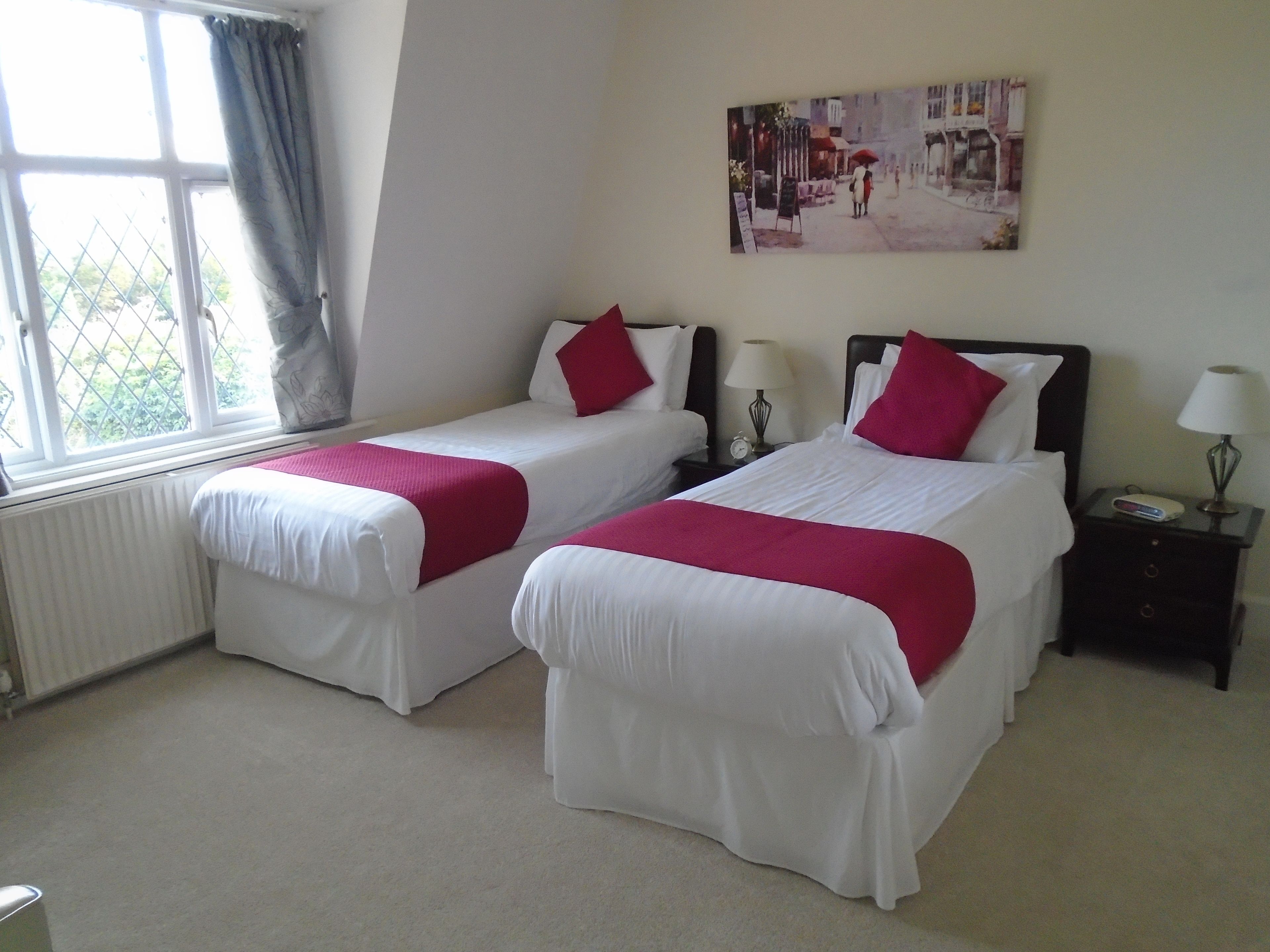 A hotel room with two single beds, a window with curtains, a wall-mounted painting, a bedside table with a lamp, and a nightstand with a clock.