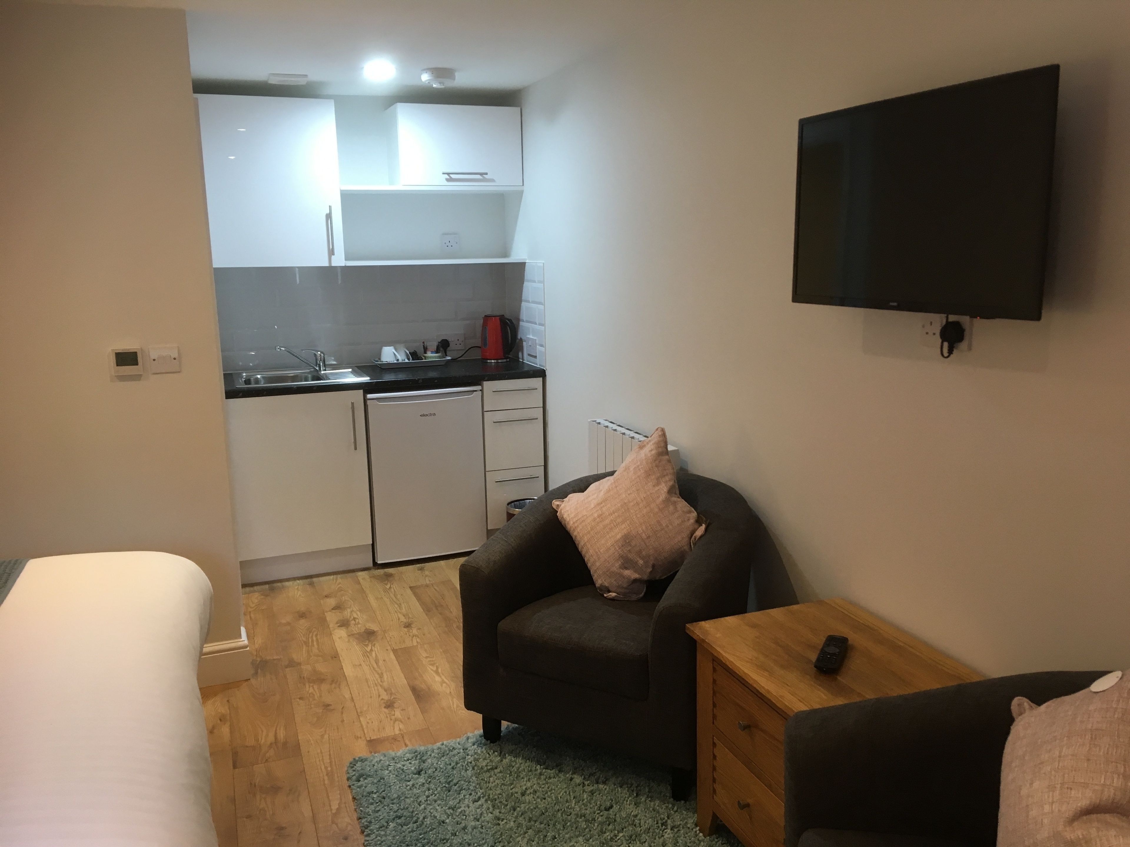 A compact living space with a kitchenette, a single bed, a small armchair, a wooden nightstand, and a flat-screen TV mounted on the wall.