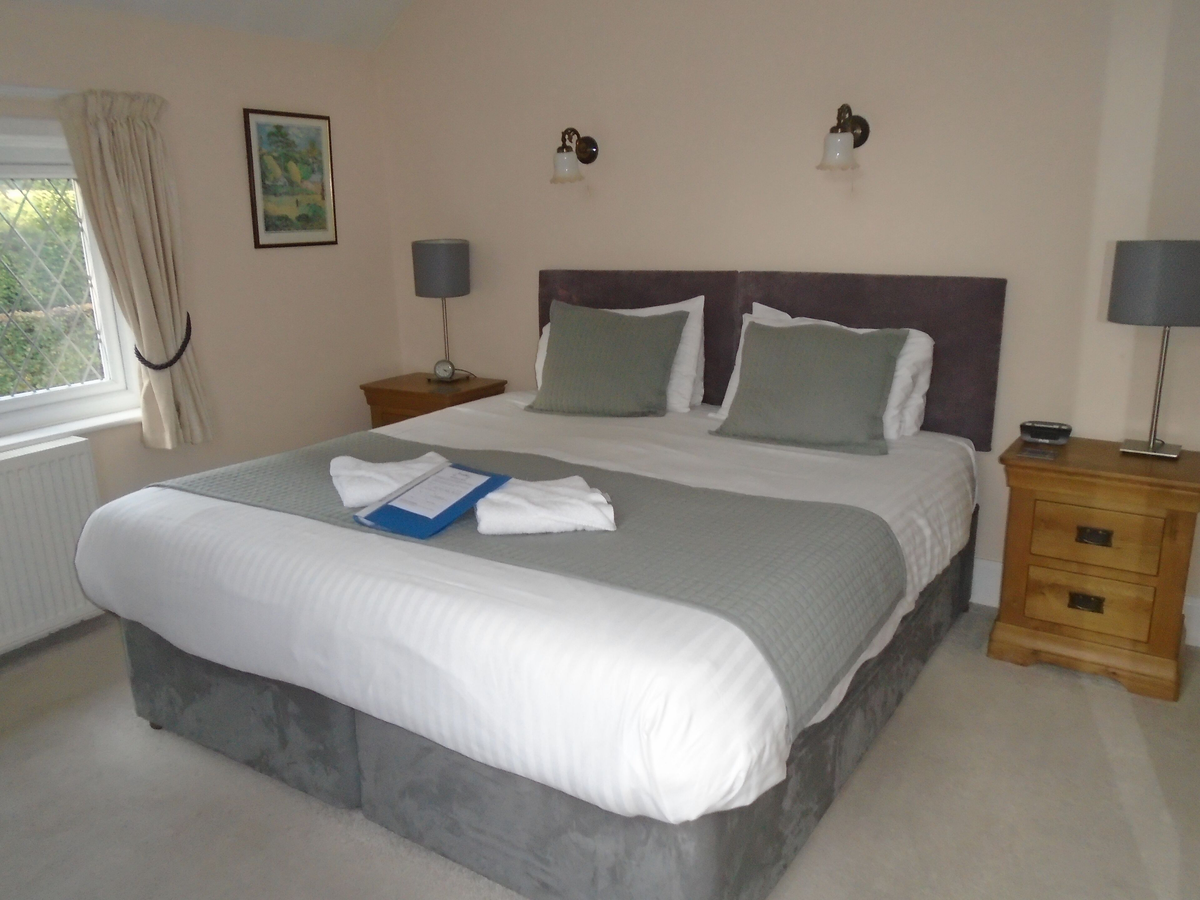 A neatly made bed with a blue folder and white towels, two bedside lamps, a nightstand with a clock, and a window with curtains.
