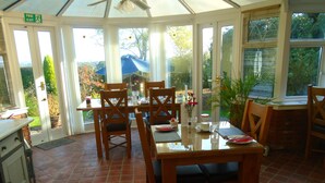 Un invernadero con una mesa de comedor dispuesta para cuatro personas, rodeada de sillas, y vistas al jardín exterior.