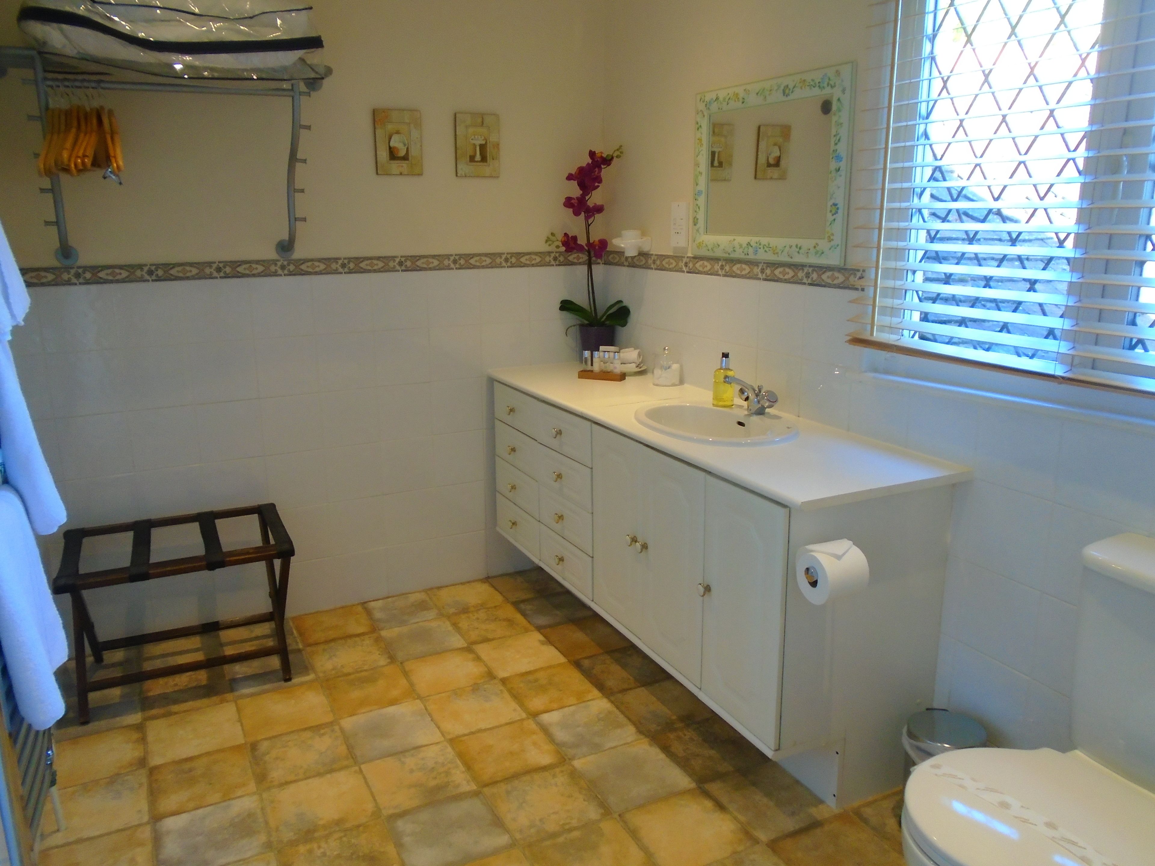 Un baño con lavamanos, espejo e inodoro blancos. Hay un estante pequeño con artículos de aseo y una planta en maceta. El suelo presenta un diseño de baldosas en tonos amarillo y marrón.