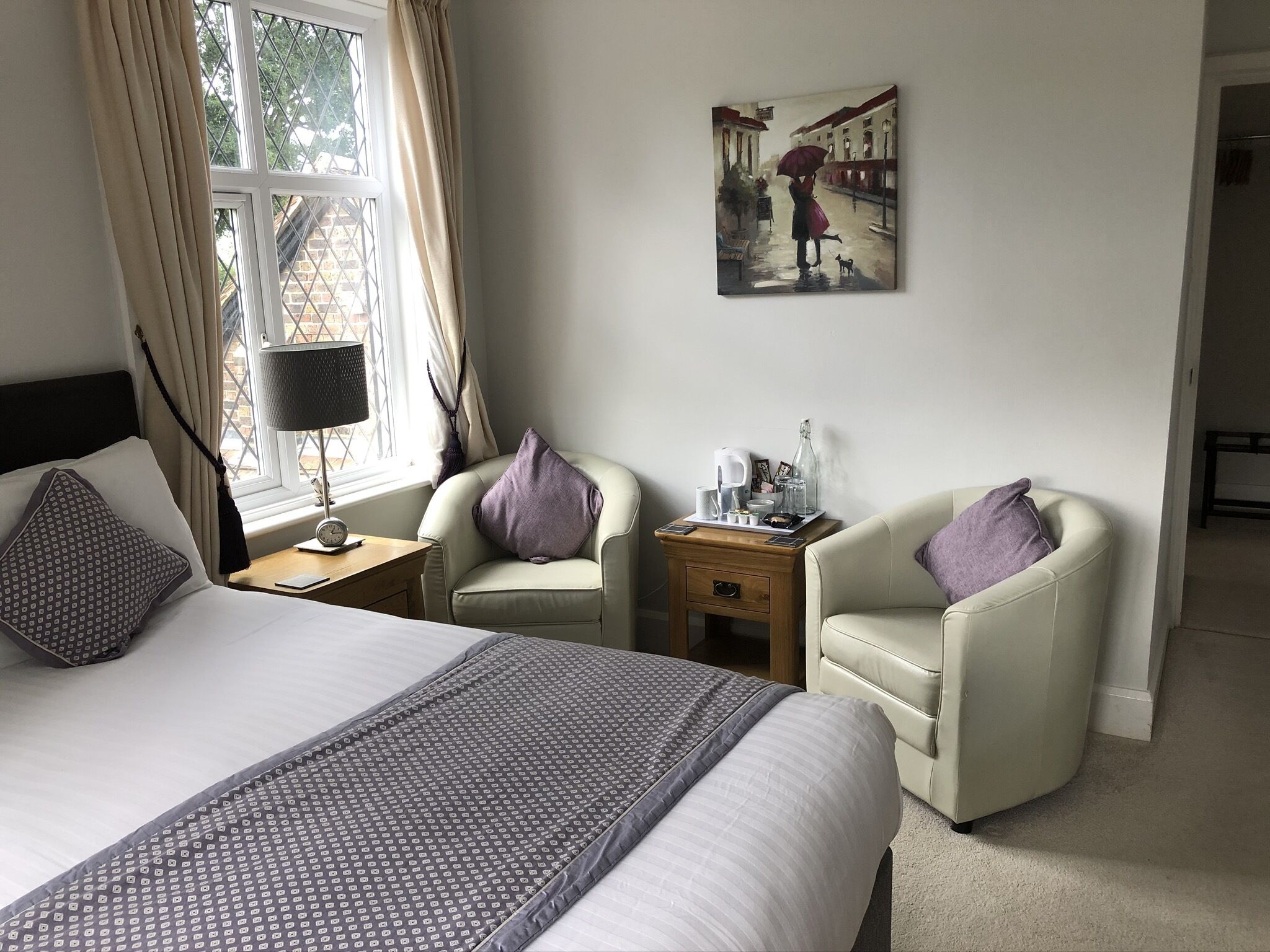 Habitación de hotel con cama, dos sillones, mesita de noche con lámpara, un cuadro en la pared y ventana con cortinas.