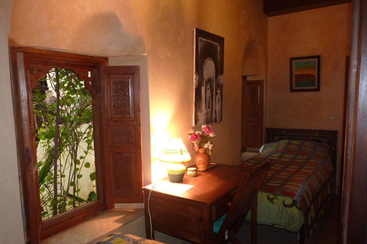Ein Schlafzimmer mit einem hölzernen Schreibtisch, einer Lampe, einer Vase mit Blumen, einem Bett mit gemustertem Überwurf und einer Tür mit dekorativer Gestaltung.