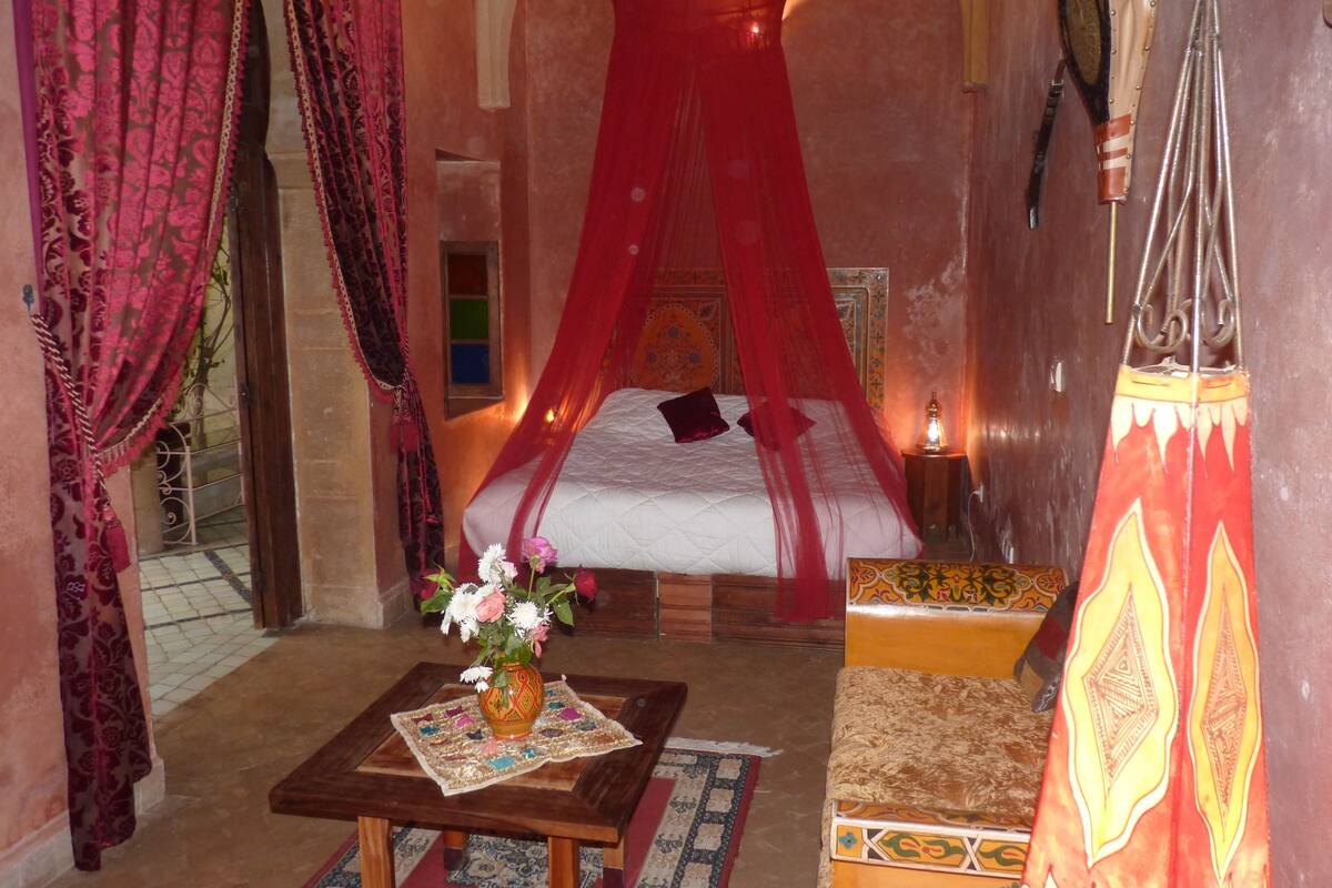 Ein Zimmer mit einem Bett unter einem roten Baldachin, einem kleinen Tisch mit Blumen und traditioneller Einrichtung.