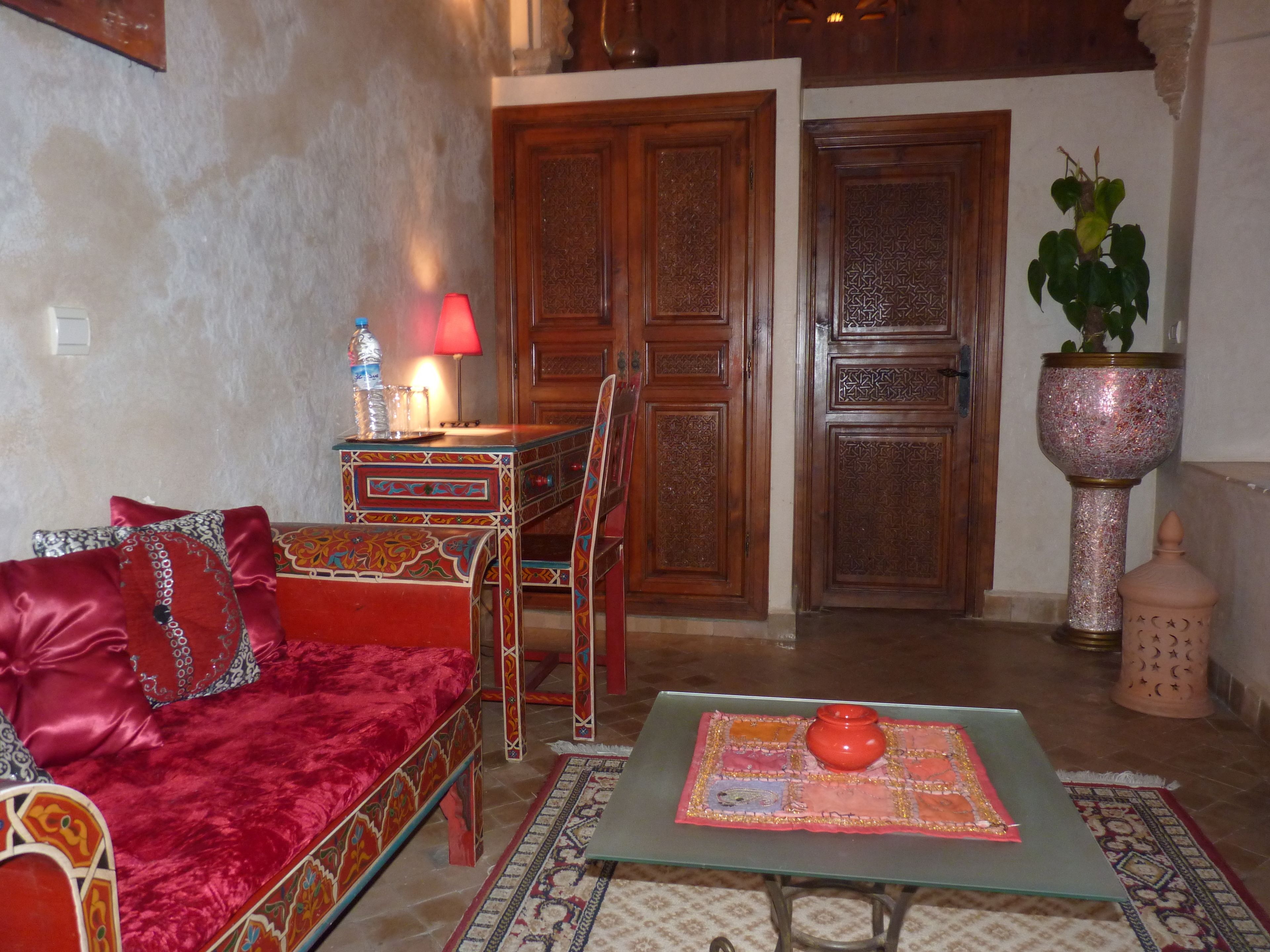 Una sala de estar acogedora con un sofá rojo, una mesa de centro de cristal y una alfombra estampada. Se ve una puerta de madera y una planta en maceta al fondo.