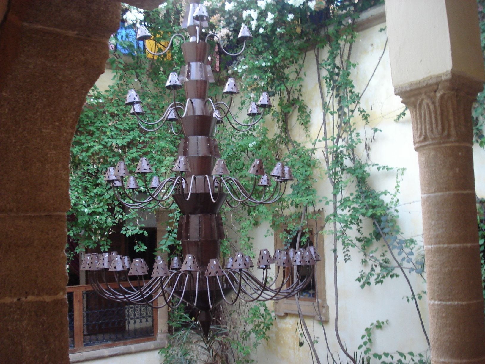 A wrought iron chandelier with multiple arms and hanging bells, set in an outdoor area with ivy-covered walls and stone columns.