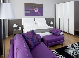 Quarto moderno com cama, duas mesas de cabeceira, um criado-mudo, uma pintura abstrata grande, um guarda-roupa e mobiliário roxo.