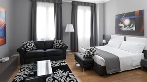 Habitación de hotel moderna con sofá de cuero negro, cama blanca, reposapiés gris, alfombra estampada y un cuadro en la pared.
