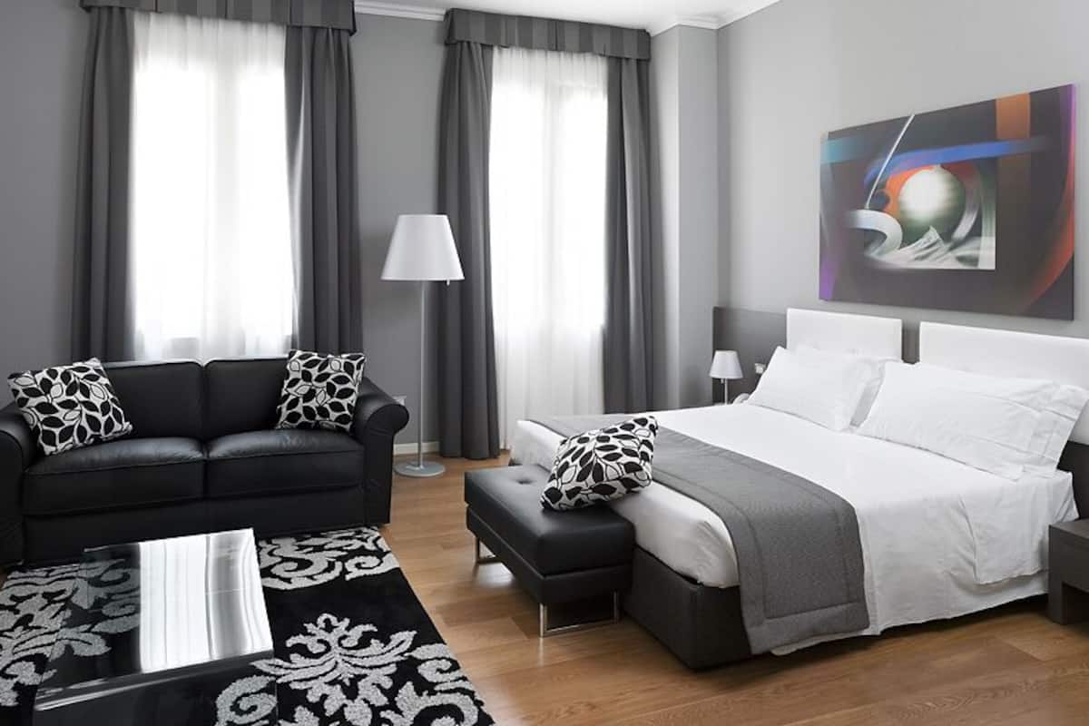 A modern hotel room with a black leather sofa, a white bed, a grey ottoman, a patterned rug, and a painting on the wall.