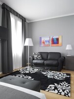 Una sala de estar moderna con un sofá de cuero negro, una mesa de centro de vidrio, un tapete con diseño blanco y negro y dos cuadros abstractos en la pared.