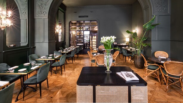 Un interior de restaurante con una mesa negra central, sillas tapizadas de verde y una zona de bar al fondo.