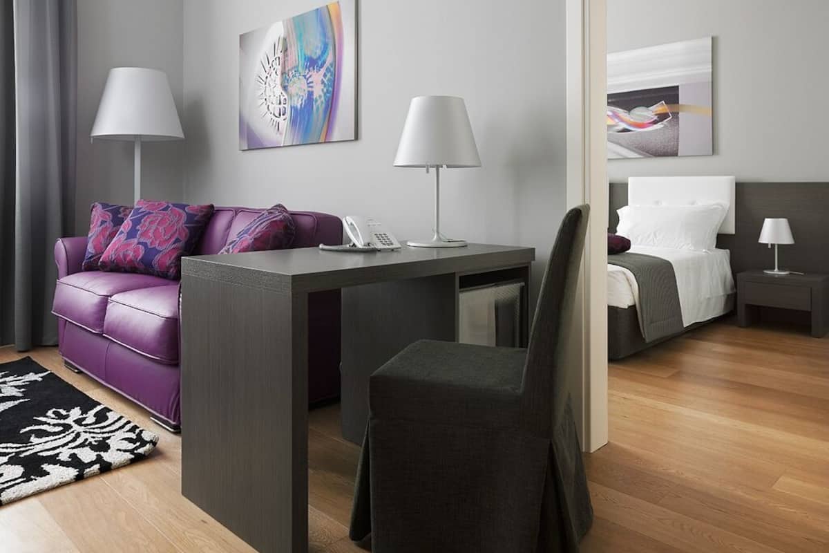 A modern hotel room with a grey sofa, a small desk with a telephone, a grey chair, a bed with white bedding, and two wall-mounted lamps.