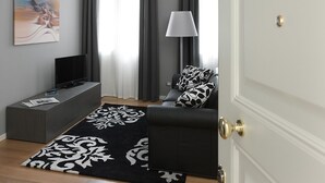 Una sala moderna con un televisor de pantalla plana, un sofá con cojines estampados, una lámpara de pie y un ventanal con cortinas grises.