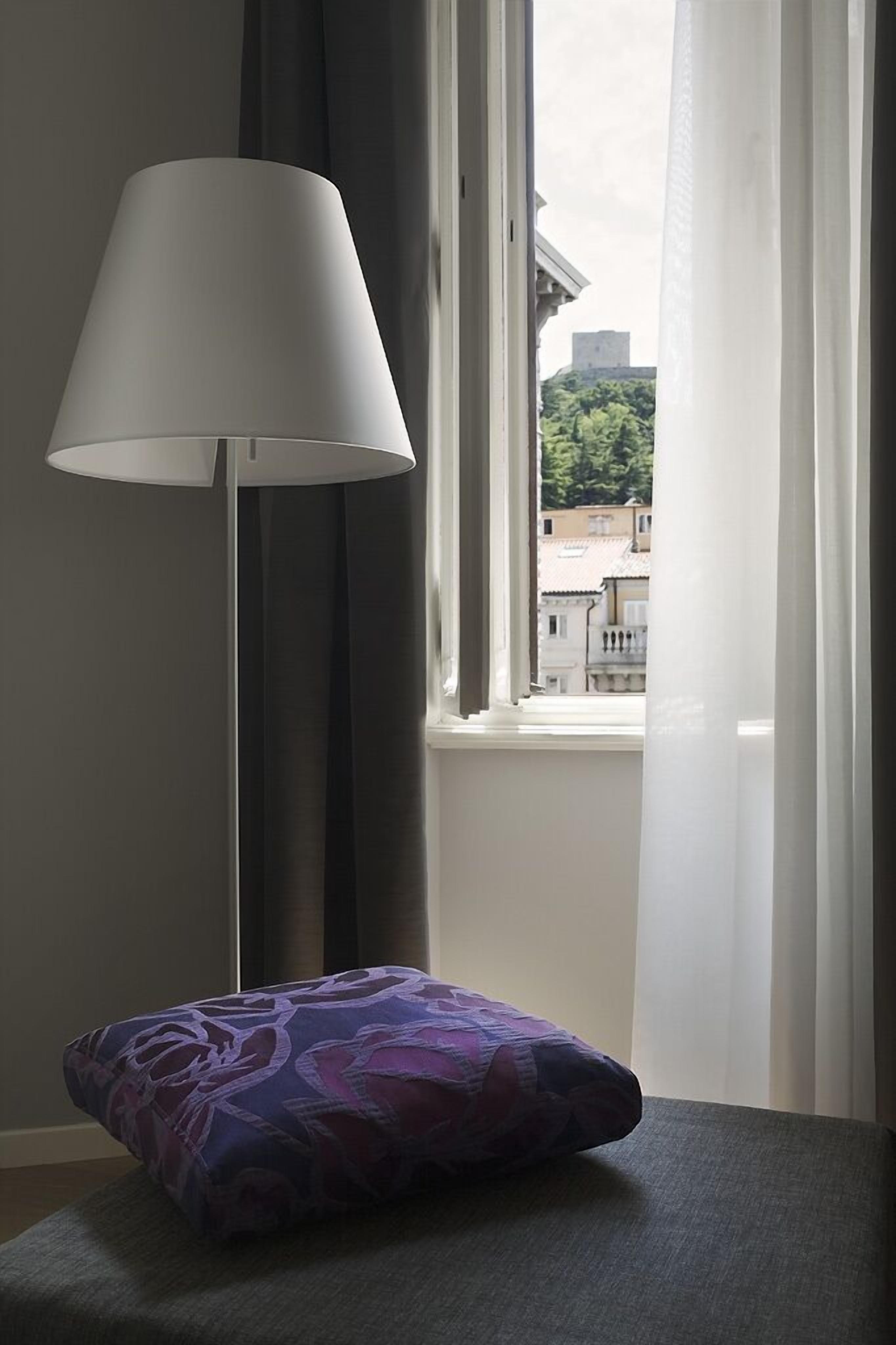 A room with a window, a floor lamp, and a cushion on a table.