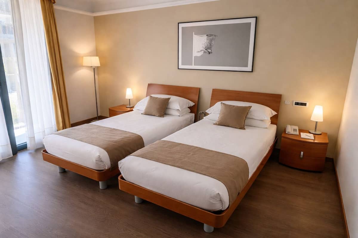 Habitación de hotel con dos camas, lámparas de noche, un cuadro en la pared y una ventana con cortinas.