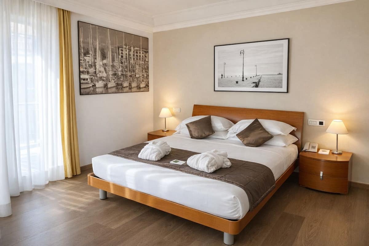 Habitación de hotel con cama, dos mesitas de noche con lámparas, un ventanal con cortinas y dos cuadros en la pared.