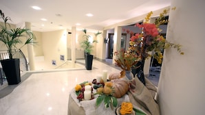 Un vestíbulo de hotel con piso de mármol, plantas en macetas, una mesa decorativa con flores y velas, y vista al interior.