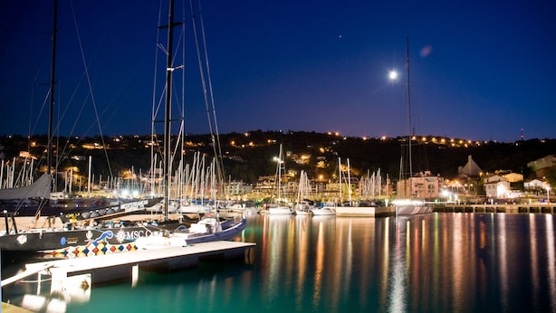 Un puerto deportivo de noche con barcos amarrados, un cielo despejado y un paisaje urbano al fondo.