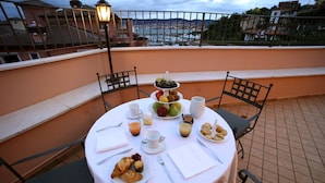 Una mesa redonda con mantel blanco, rodeada de cuatro sillas, ubicada en un balcón con vistas a un paisaje urbano que incluye edificios y una masa de agua.