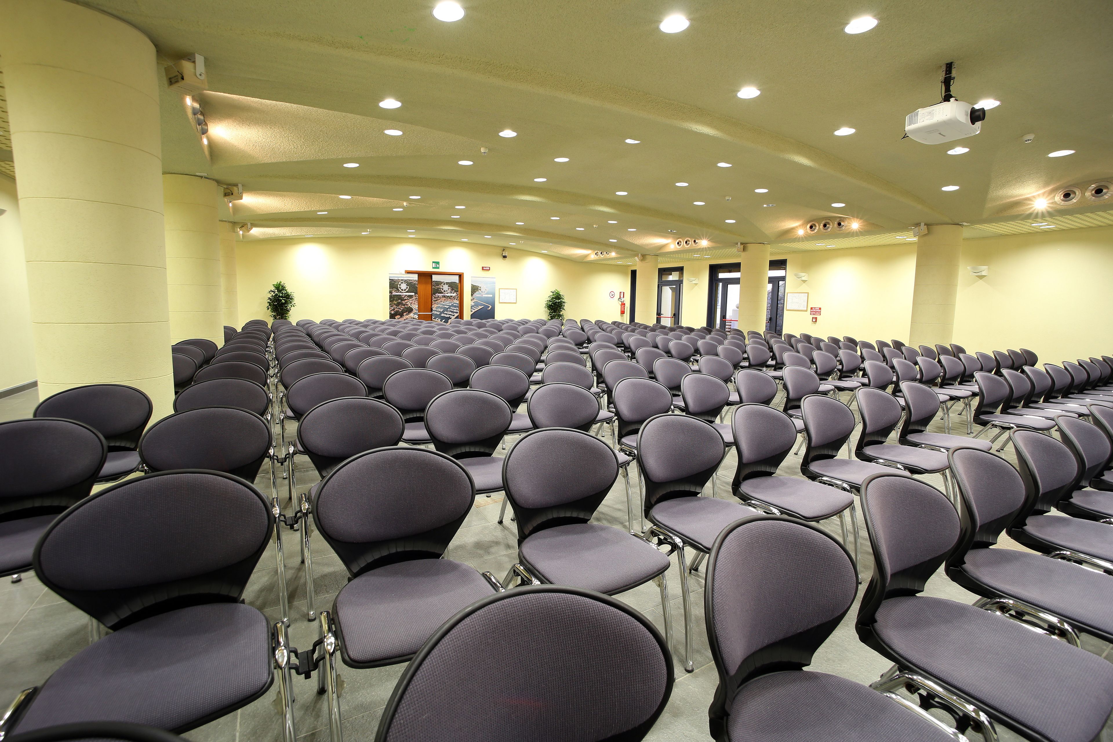 Una sala conferenze con file di sedie, un proiettore e una porta sullo sfondo.