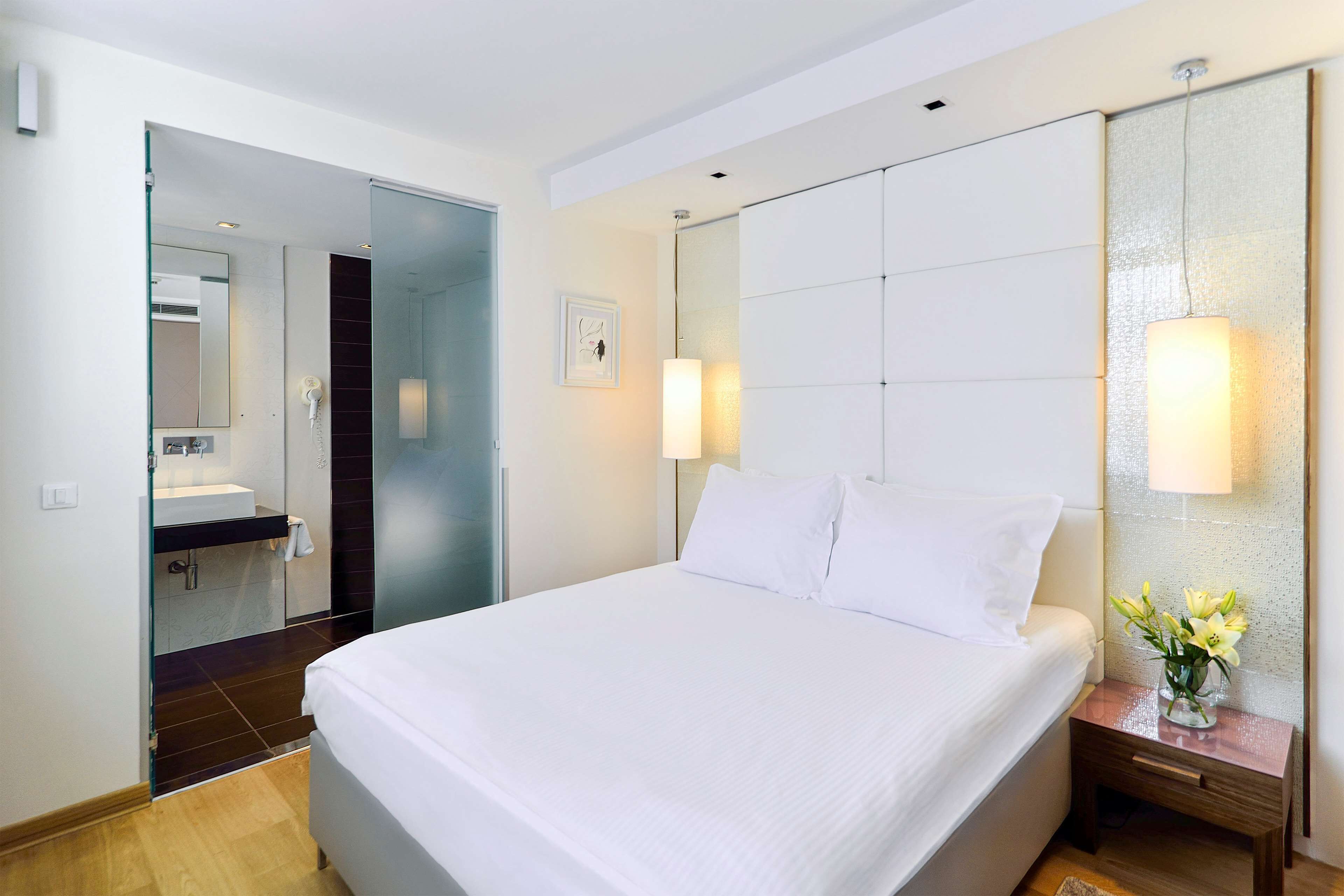 Quarto de hotel moderno com uma cama grande, uma mesa de cabeceira com abajur, um espelho e um banheiro visível por uma porta aberta.