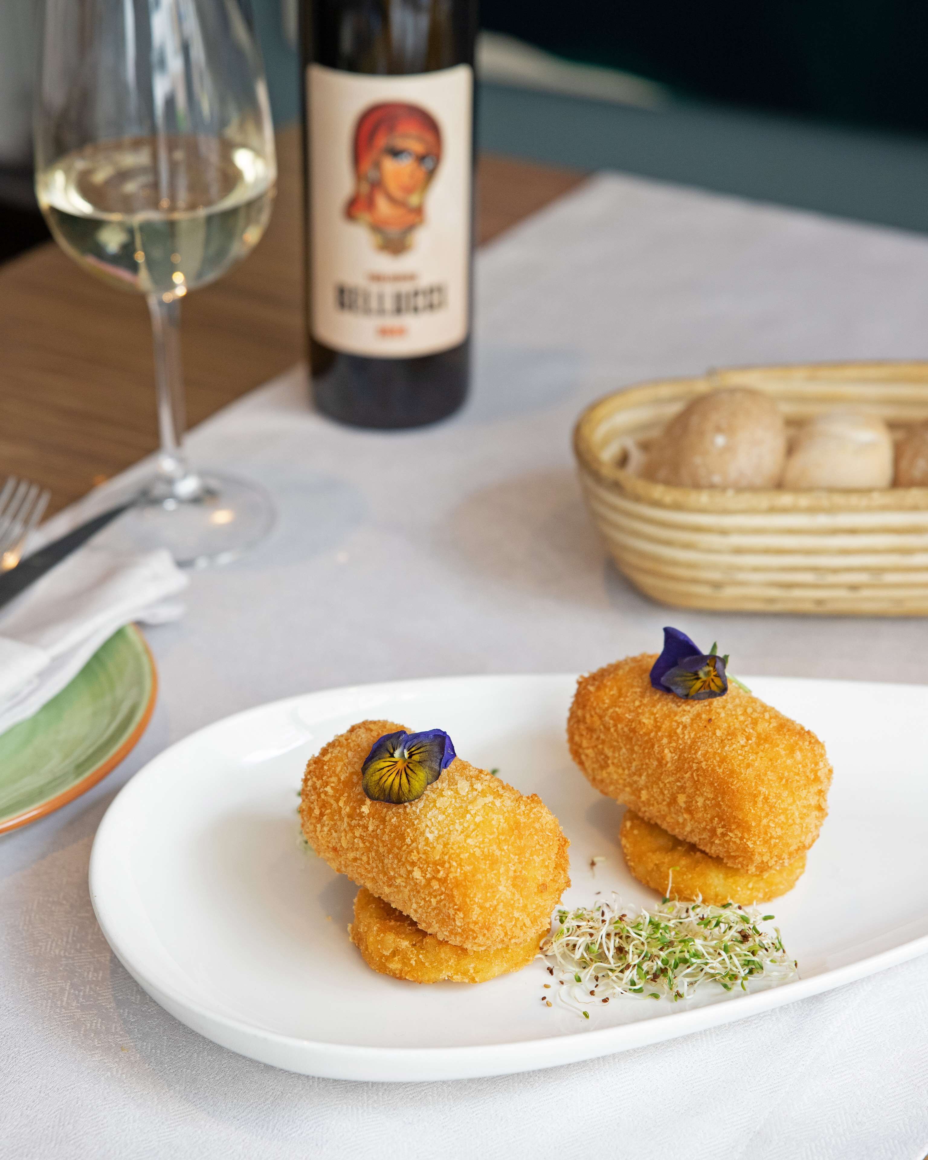 Un plato blanco con dos panes empanizados adornados con una flor, una botella de vino y una cesta de bollos sobre una mesa.