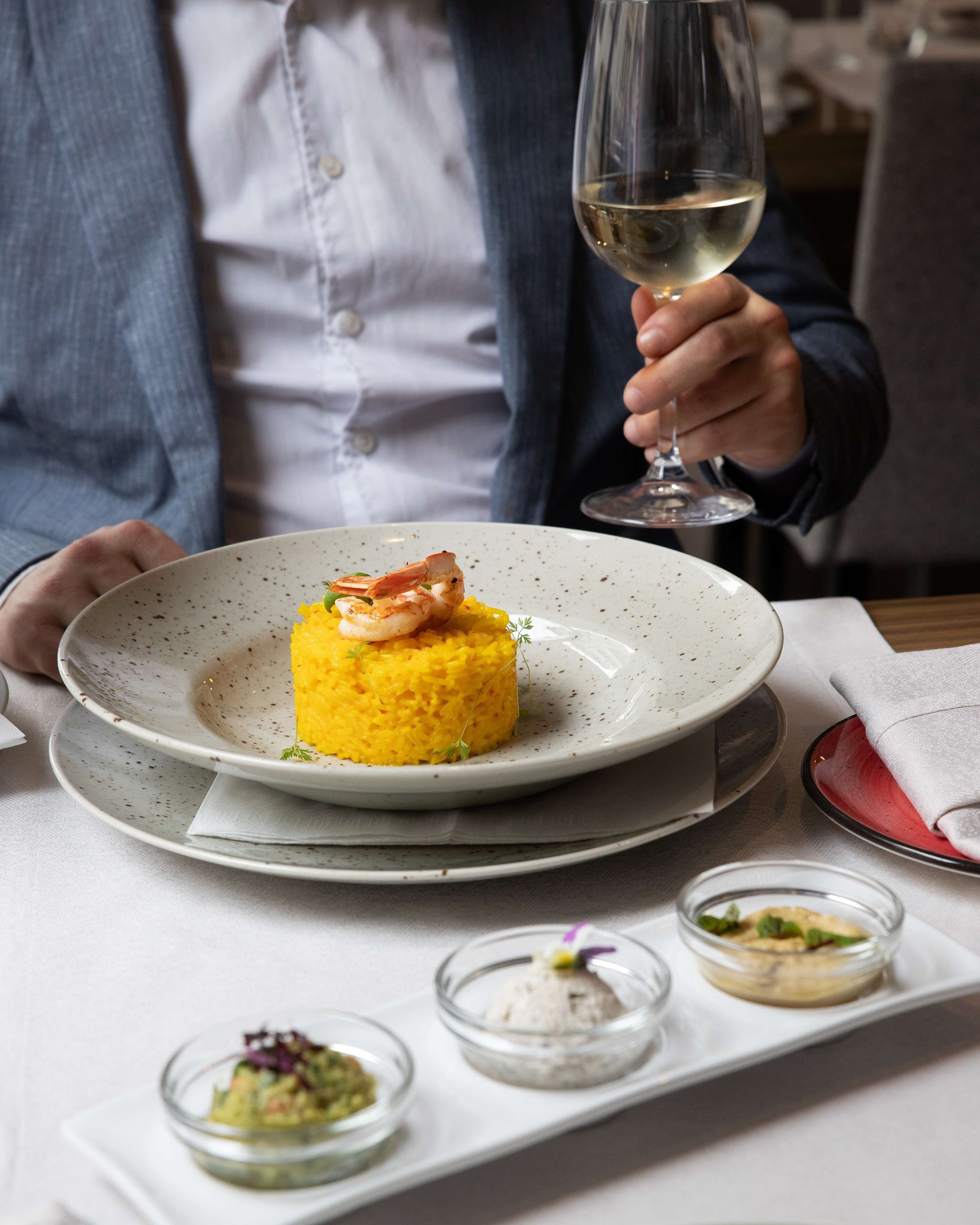 A person holding a wine glass, seated at a table with a plate of food, a napkin, and small bowls of condiments.