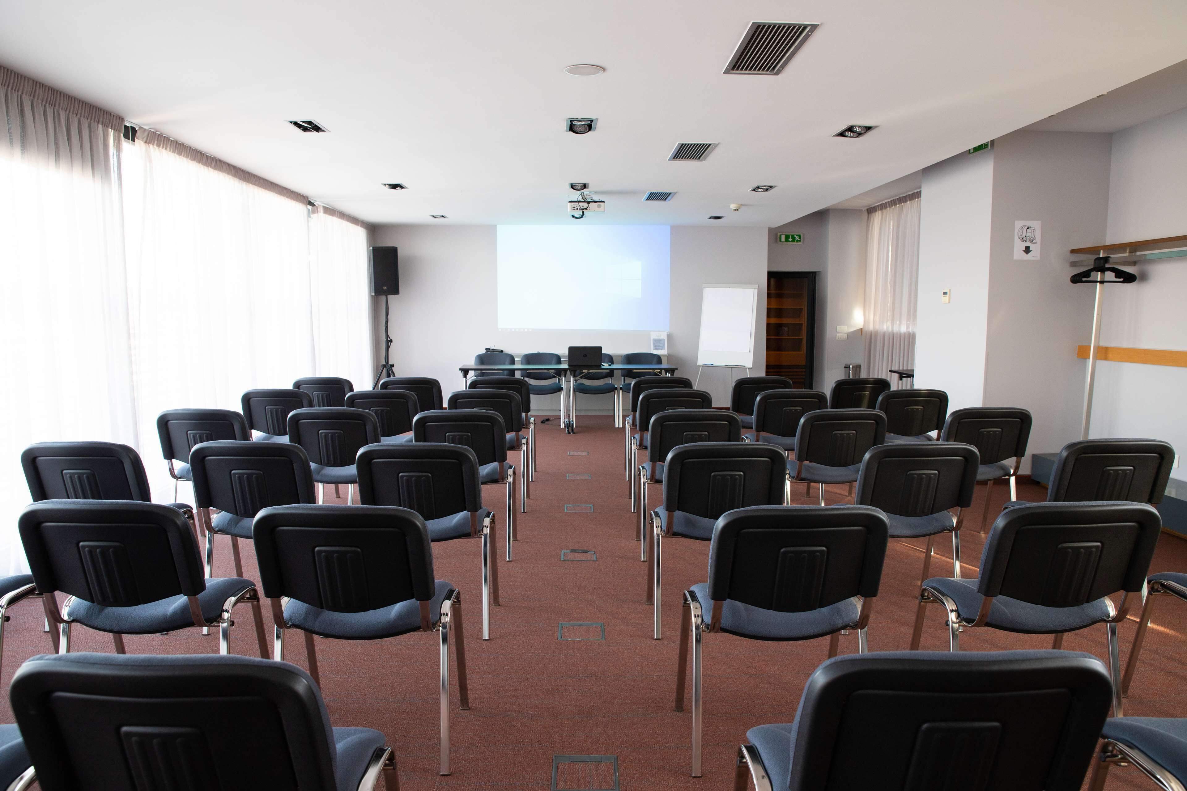 A conference room with rows of chairs, a projector screen, a whiteboard, and a flat-screen TV.