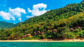 Una zona costera con playa y una colina con varias construcciones pequeñas de techo de paja.
