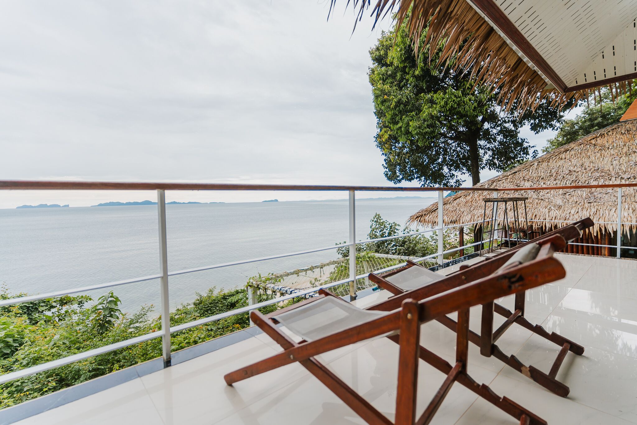 A balcony with a view of the sea, wooden chairs, and a thatched roof structure.