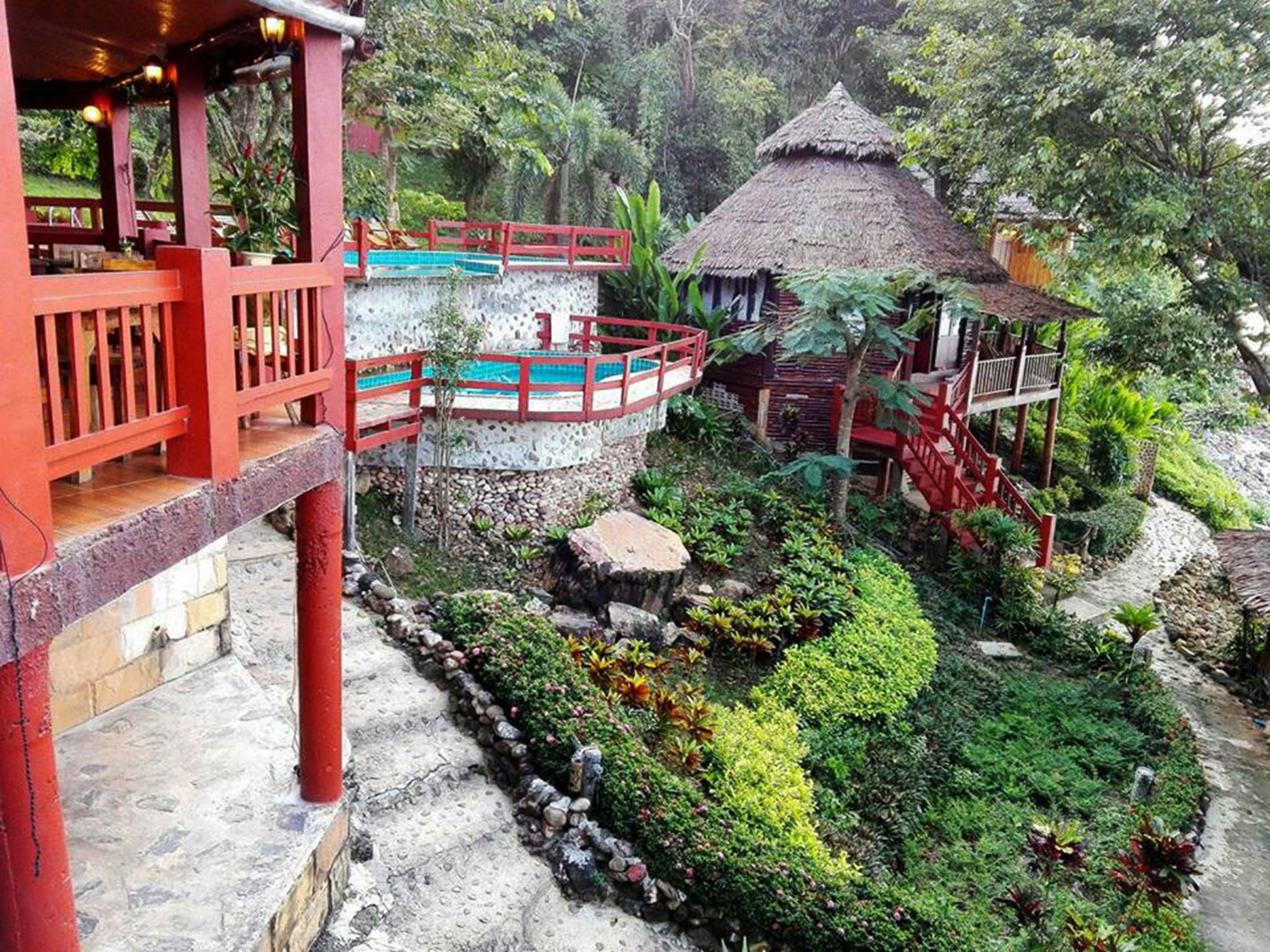 A tropical resort with a pool, a thatched-roof hut, and a red-roofed structure with a balcony.