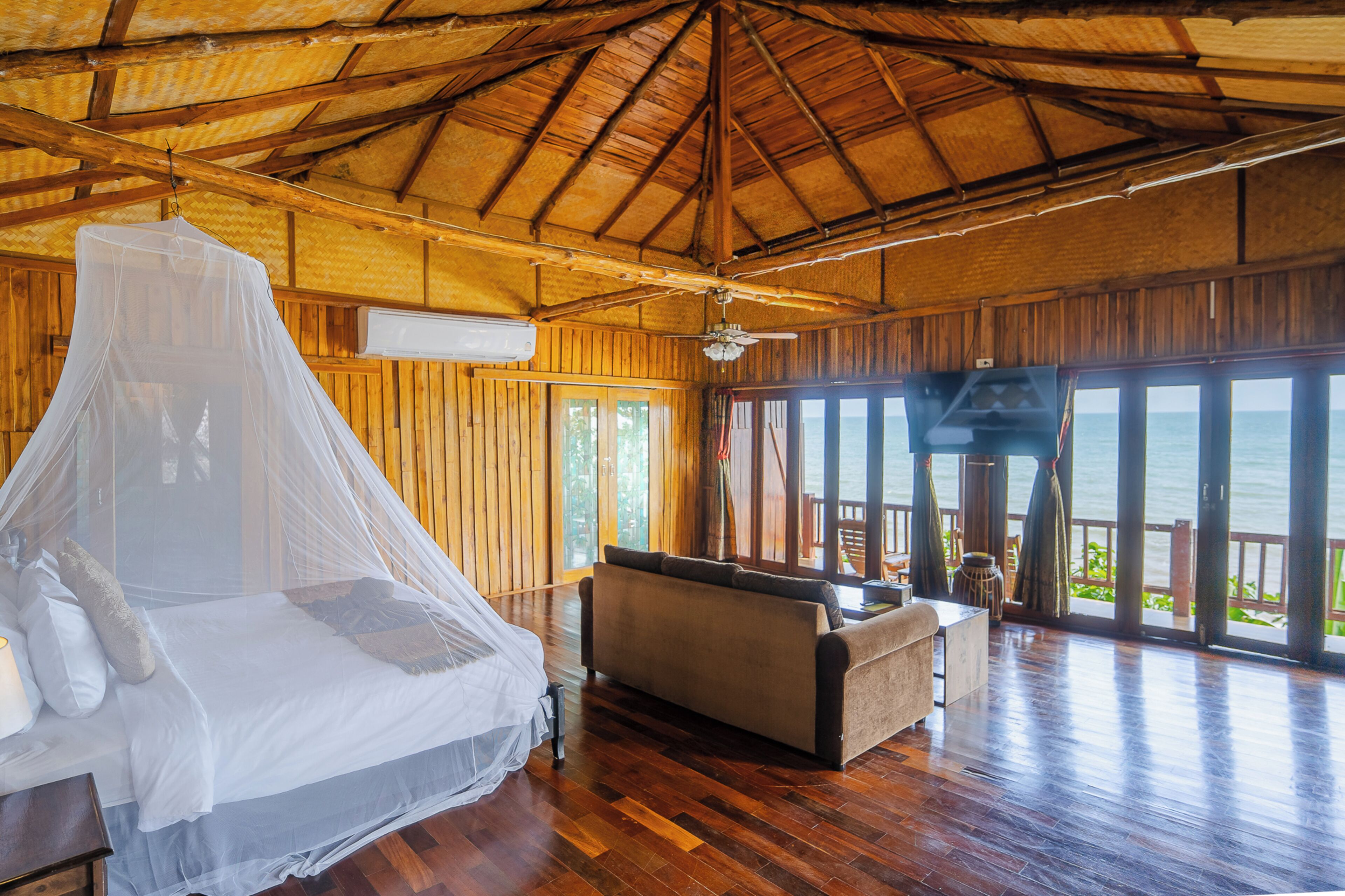 A room with a bed under a mosquito net, a sofa, and large windows overlooking the sea.