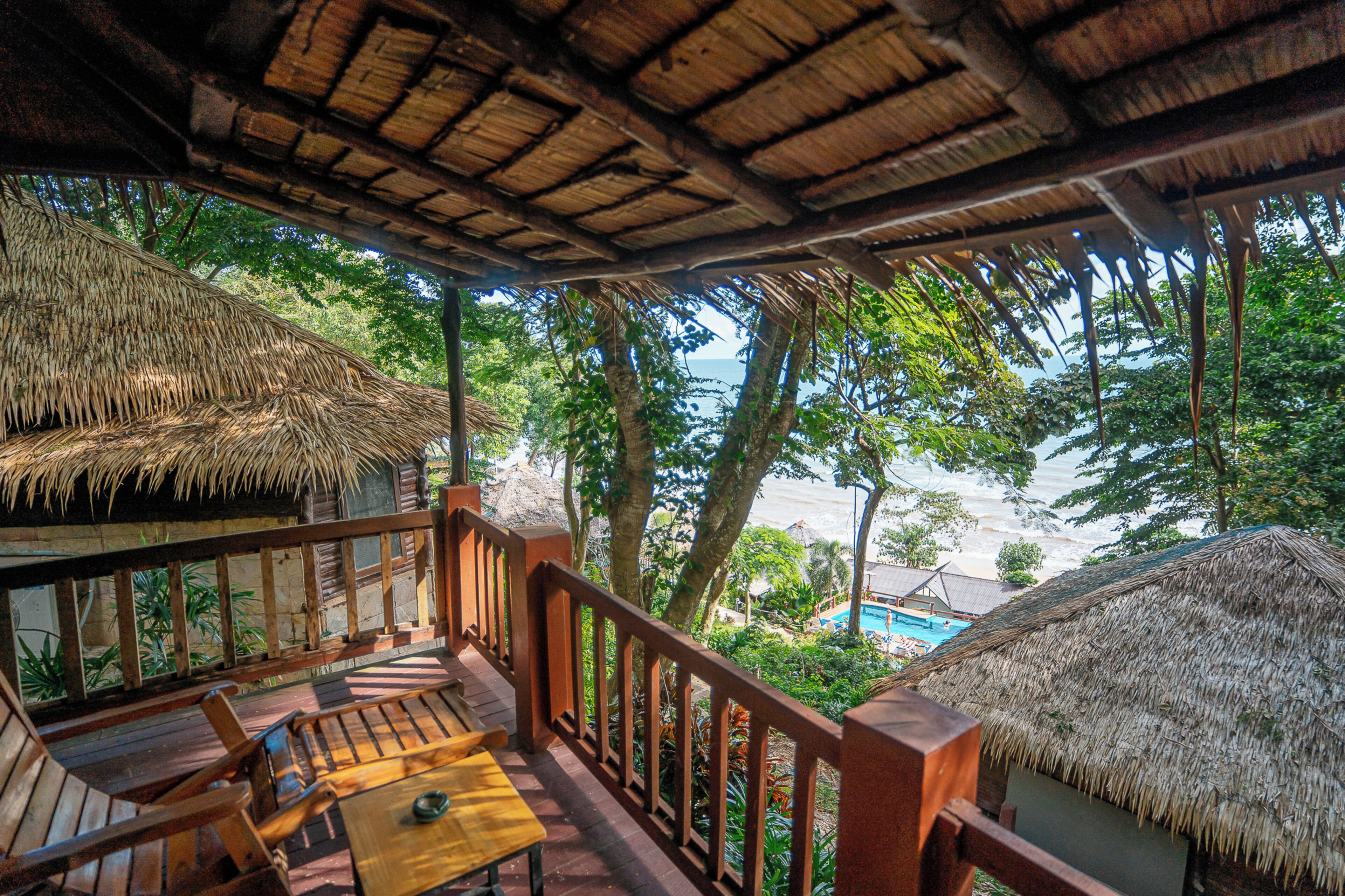 Une terrasse en bois surmontée d’un toit de chaume, offrant une vue sur une plage et une végétation luxuriante.