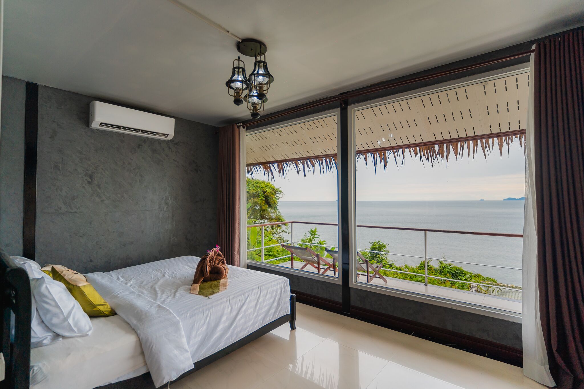 A bedroom with a bed, an air conditioner, a chandelier, a large window with a view of the sea, and a balcony with chairs.