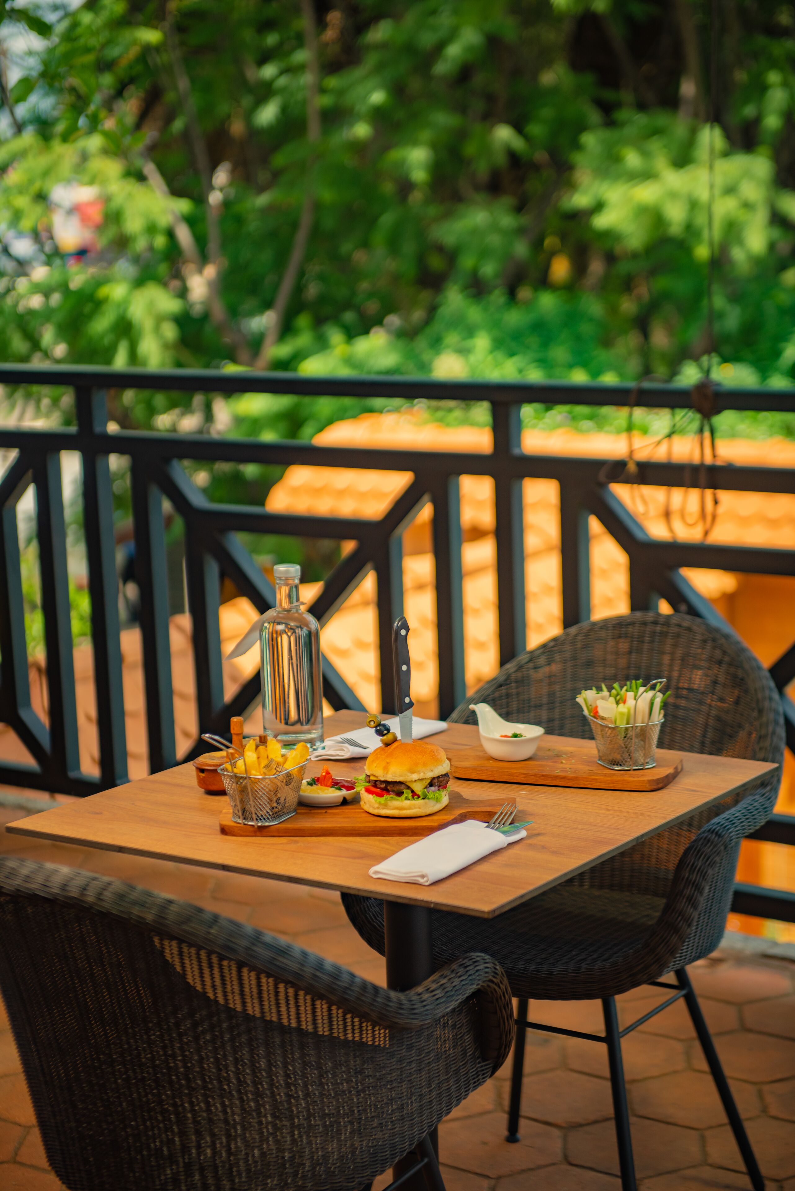 A wooden table with a bottle, a knife, a basket of fruit, a burger, and a small bowl of salad on a patio with wicker chairs and a railing.