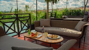 A terrace with wicker furniture, a wooden table with food and drinks, and a view of greenery and buildings. - Radisson Hotel Tamboho Waterfront Antananarivo (Antananarivo)