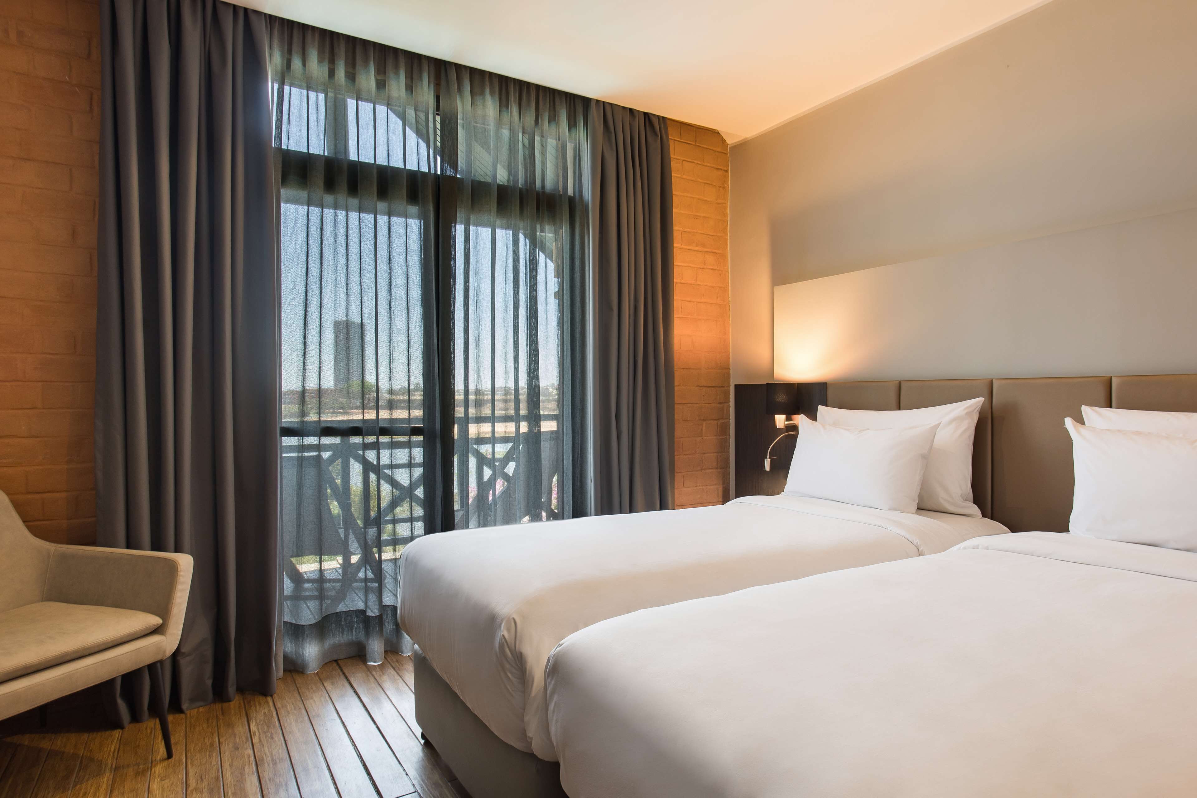 A hotel room with two beds, a chair, a window with curtains, and a view of the outdoors.