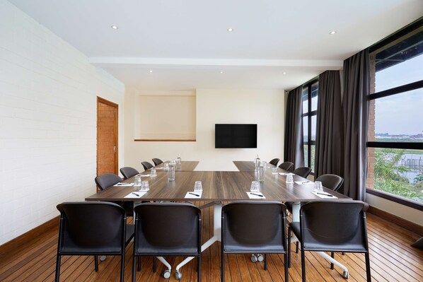 Une salle de conférence avec une longue table, des chaises noires, un téléviseur à écran plat et de grandes fenêtres équipées de rideaux.