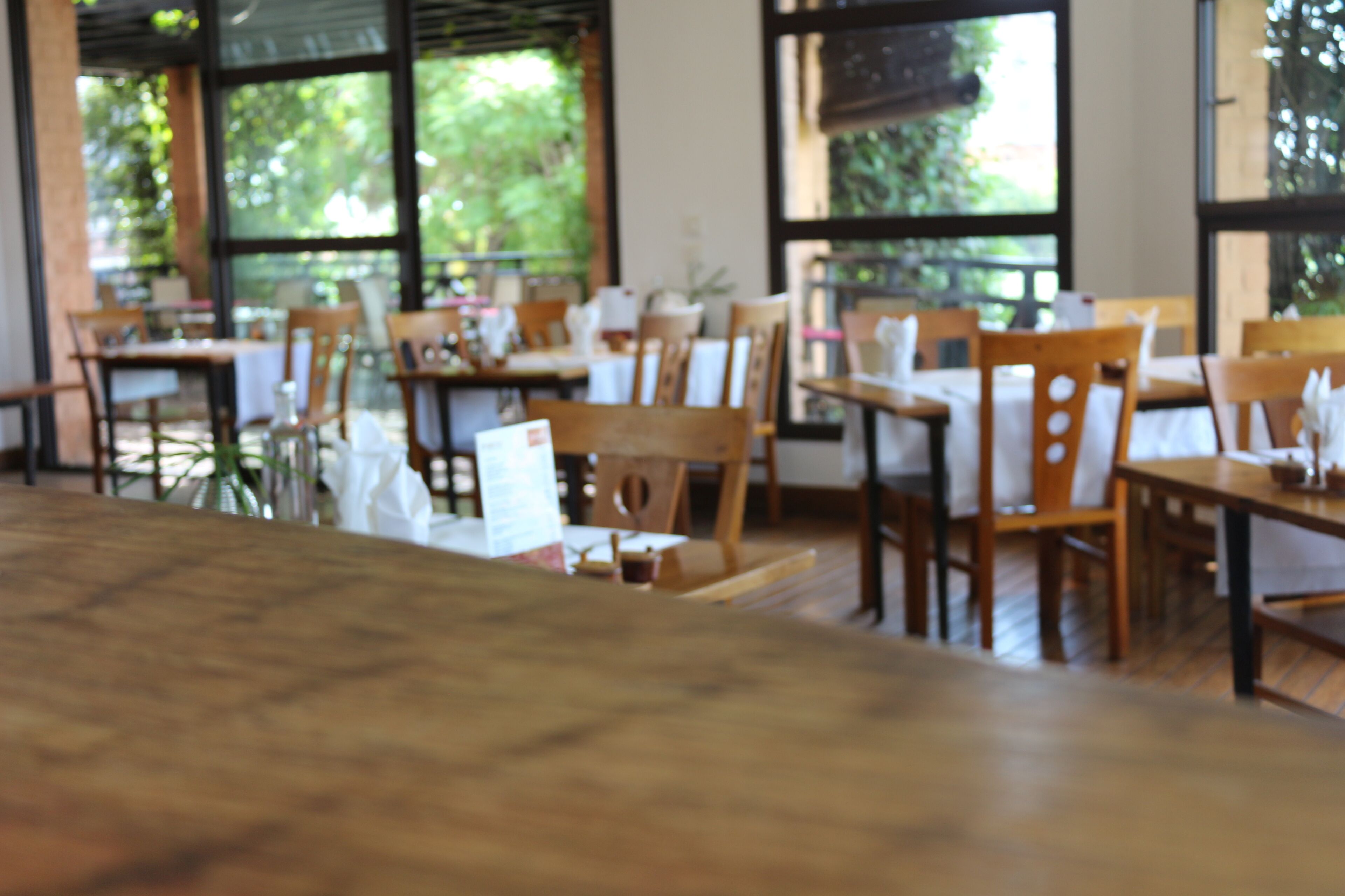 A restaurant with wooden tables and chairs, large windows with a view of greenery outside.