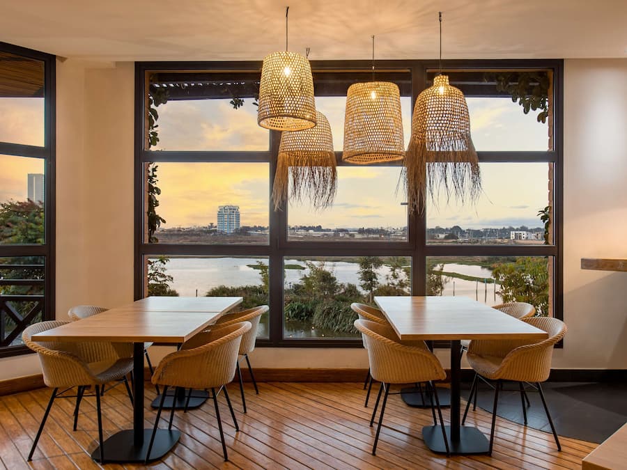 Un comedor con un ventanal grande, sillas de mimbre, una mesa de madera y lámparas colgantes de ratán.