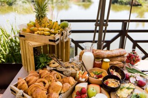 A breakfast buffet with fruits, pastries, and beverages on a wooden table by a lakeside. - Radisson Hotel Tamboho Waterfront Antananarivo (Antananarivo)