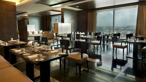 Un comedor moderno de hotel con mesas dispuestas para comer, amplias ventanas que ofrecen vista a la ciudad y un diseño interior contemporáneo.